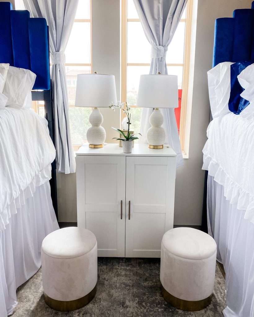 a dorm room with two beds on either side with stools on the floor. a white side table is in the middle with two white lamps resting on it. the bed has navy blue headboards to bring in pops of blue.
